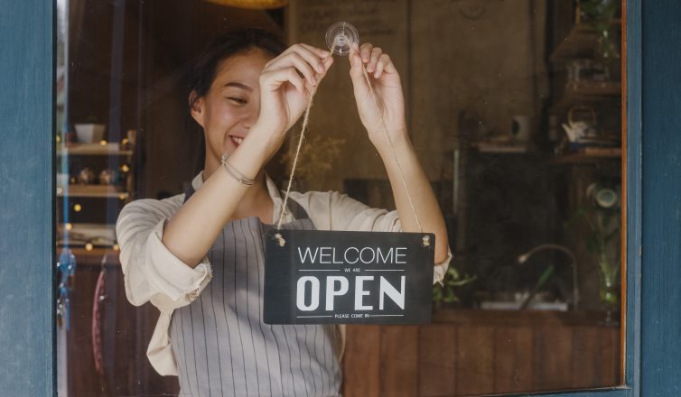 Woman hanging a “welcome, we’re open” sign on the glass door of a store
